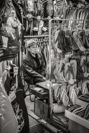 Black and white portrait of a Tokyo shopkeeper beside racks of sandals