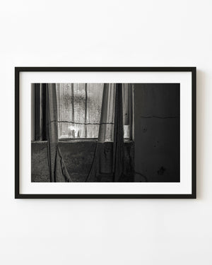 Torn curtain lit by a window beside a cracked wall in black and white