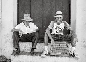 Black and white Trinidad street portrait of two seated men with cigars and a birdcage.