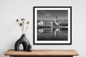 Square monochrome photograph of Trogir architecture, a low bridge and water reflections.