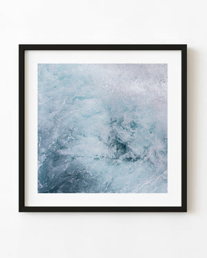 White sea foam churns through pale blue water in a square abstract photograph.