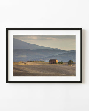 Tuscan farmhouse and cypress trees amid plowed fields beneath layered hills.