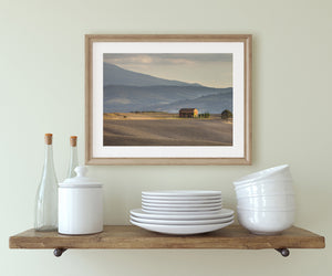 A Tuscan farmhouse stands beside slender cypress trees with hazy hills beyond.