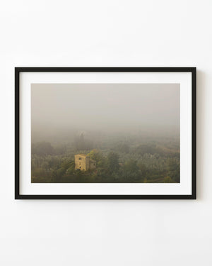 Tuscan stone farmhouse surrounded by olive trees beneath thick morning fog.