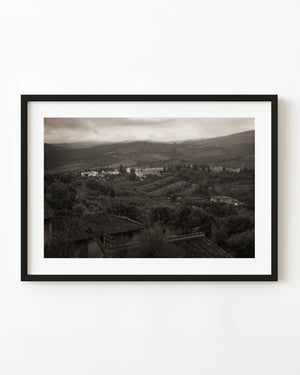 Tuscan village and rolling countryside beneath a cloudy sky in warm monochrome.