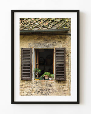Open Tuscan window with dark wooden shutters and potted plants on a stone wall.