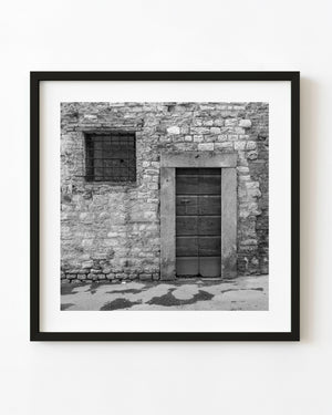 Weathered Tuscan wooden door with a small cross beside a barred window in black and white.