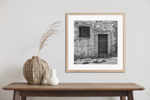 Square monochrome photograph of rustic masonry, a timber doorway and damp pavement.