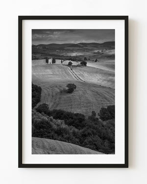 Small Tuscan chapel and farmhouse among rolling fields in black and white.
