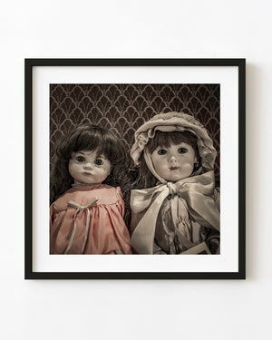 Two antique porcelain dolls in softly toned clothing against a patterned backdrop.