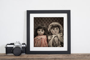 Square still-life photograph of two antique dolls in pink and cream dresses.