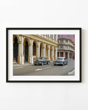 Two classic cars pass a yellow colonnaded building on a Havana street.