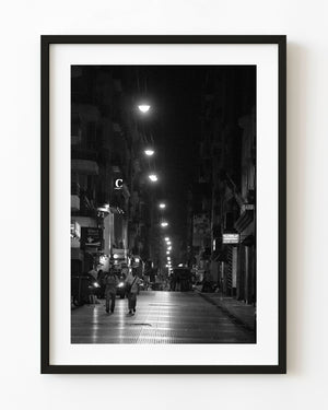 Two men walk beneath a receding line of streetlights in a black and white night photograph.