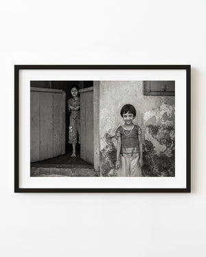Two sisters beside a weathered doorway in India, photographed in black and white.