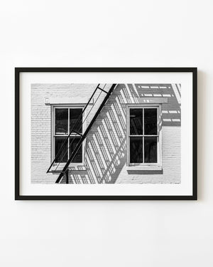 Two windows and a diagonal fire escape cast sharp shadows on a brick wall in black and white.