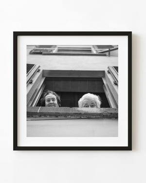 Two older women look down from an open Tuscan window in black and white.
