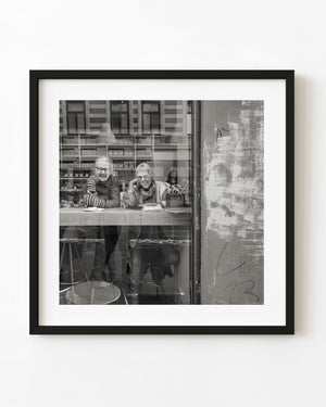 Two women laugh together at an Oslo cafe window in black and white.