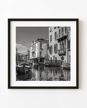 Venetian canal homes and moored boats reflected in rippling water in black and white