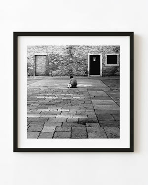 Boy crouching beside a football on a paved Venice square in black and white