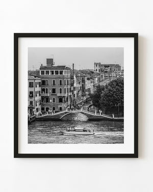 Venice canal bridge with pedestrians and a passing motorboat in black and white