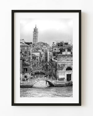 Venice canal bridges and a tall bell tower above tiled rooftops in black and white