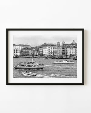 Boats cross a busy Venice waterfront beneath historic buildings in black and white