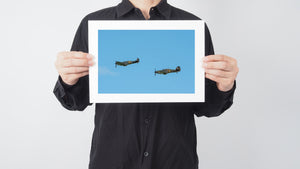 Two camouflaged propeller planes crossing a pale blue sky.