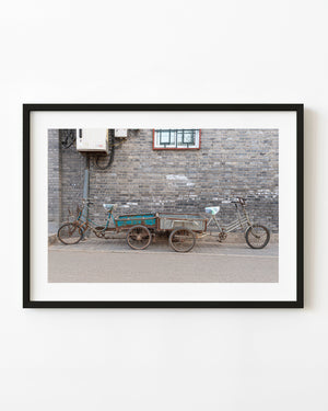 Two weathered cargo tricycles against a grey brick wall in Beijing