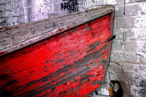 Peeling red paint on a wooden rowboat beside textured brickwork