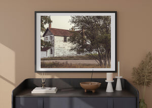 White farmhouse with worn paint and brick chimneys framed by leafy branches