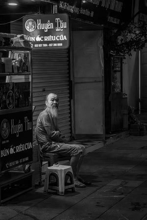 Elderly Vietnamese man seated beside a Hanoi street food cart