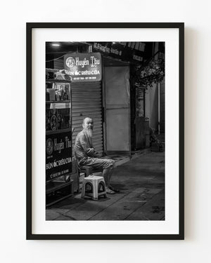 Elderly bearded man seated on a Hanoi sidewalk in black and white B