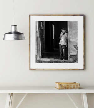 Older man wearing a brimmed hat framed by worn plaster walls