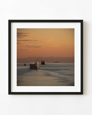 Cargo ships crossing quiet Japanese waters beneath an amber sunset sky and distant hazy mountains.