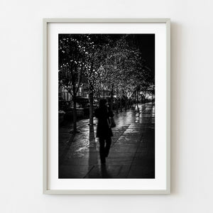 Black and white photograph of a woman walking on a wet Manhattan sidewalk at night
