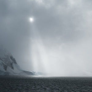 Square colour photograph of soft sunlight descending through cloud over Antarctica.