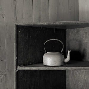 Square monochrome still life of a pale metal teapot against dark wooden boards.