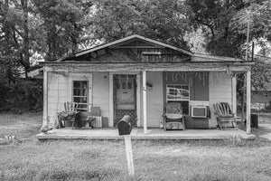 A broad front porch sits beneath a low pitched roof with trees rising behind the small house.