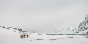 Small hikers traverse a snowy Antarctic shore with dark rocks and cloud-covered cliffs.