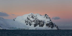Sharp Antarctic peaks and broad snowy slopes between pastel sky and rippled ocean water.