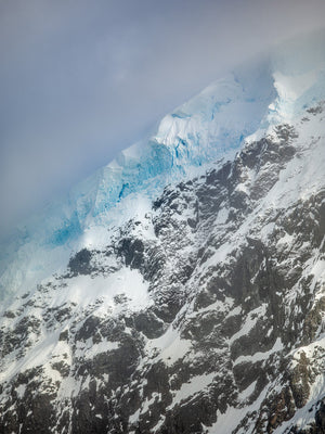 Antarctic mountain face with blue glacier ridges, dark rock and snow under low cloud.