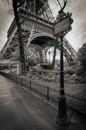 Pale street-sign lettering contrasts with dark decorative ironwork beneath the Eiffel Tower in Paris.