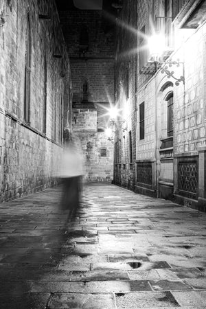 Bright wall lamps reflect across wet paving beside a motion-blurred person in Barcelona.