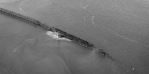 A long industrial trestle cuts diagonally across softly rippled water in monochrome.