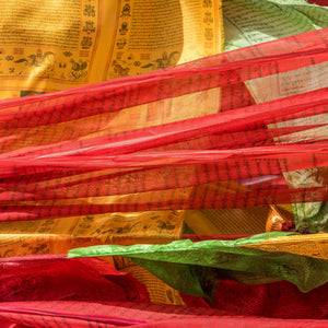 Layers of colourful prayer flag cloth overlap in a textured red and gold pattern.