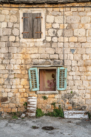 A vertical colour photograph shows two windows in a pale stone wall with blue-green shutters below.