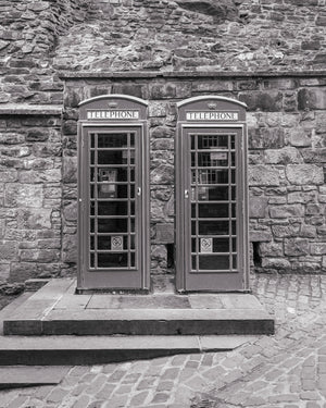 Black and white photograph of paired telephone boxes beside weathered stonework.