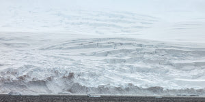A wide Antarctic landscape of layered white snow, broken ice faces and dark sea.