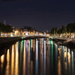Golden, green and blue city lights reflect beneath Ha’penny Bridge on Dublin’s dark River Liffey.