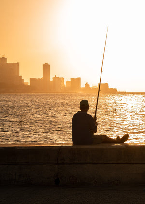 A seated fisherman holds a long rod against sunset reflections along Havana’s Malecon.